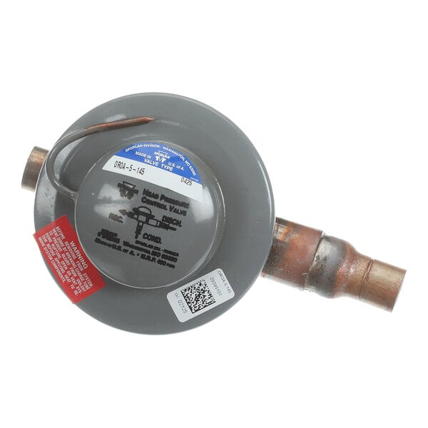 A round, gray metal valve with attached copper pipes and several labels, including a blue label, a red warning label, and a QR code.