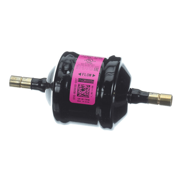 A cylindrical black component with brass fittings on both ends and a pink label in the center, indicating flow direction.