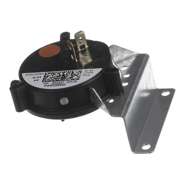 A round black component with two metal terminals and a metal mounting bracket attached, featuring a label with a QR code and some printed information.