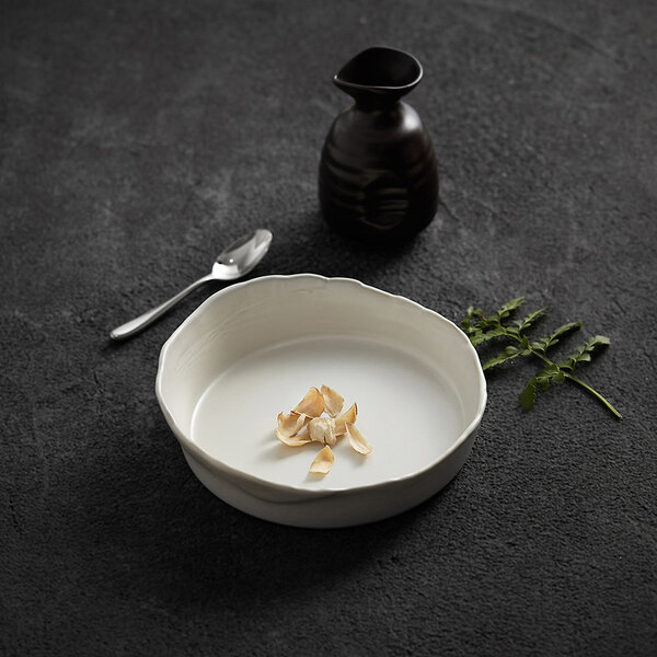 A white, round deep plate with a small pile of thin, light-colored food slices in the center, placed on a dark textured surface alongside a spoon, a dark vessel, and a sprig of green herbs.
