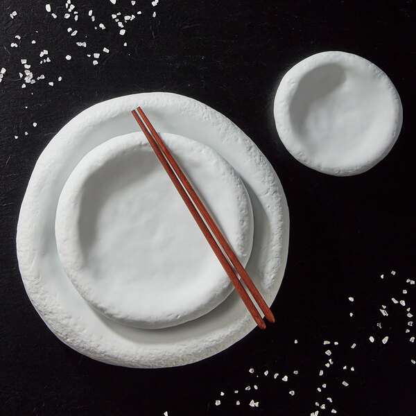 A set of three white, irregularly shaped porcelain plates with a pair of wooden chopsticks, arranged on a dark surface with scattered white granules.