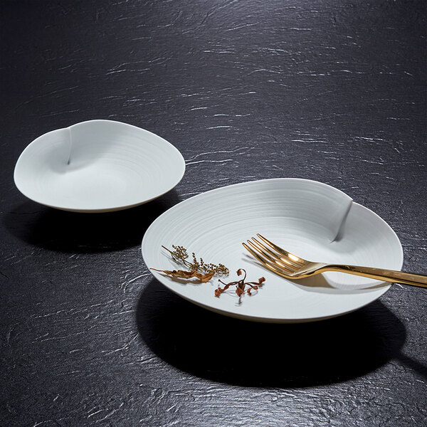 Two white, uniquely shaped porcelain plates are displayed on a dark textured surface, with one plate holding a gold fork and some dried herbs.