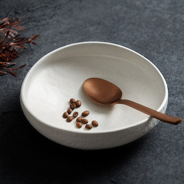 A white, textured deep plate with a bronze-colored spoon and several coffee beans inside, placed on a dark surface.