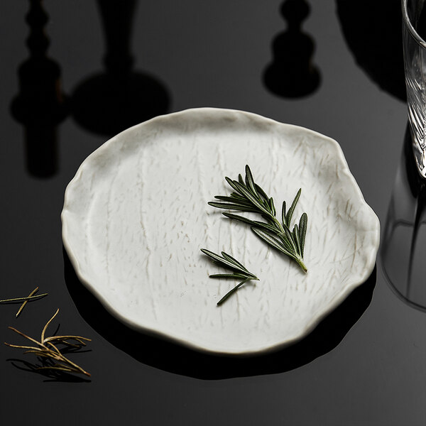 A small, irregularly shaped white plate with a textured surface, holding a sprig of rosemary, placed on a reflective black surface.