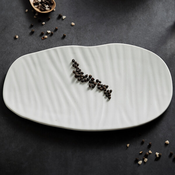 A white, oval-shaped plate with a wavy textured surface, holding a small line of whole black peppercorns.
