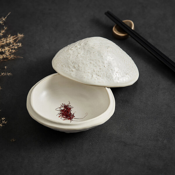 A small white stoneware bowl with a textured lid, containing a small amount of red saffron-like threads, placed on a dark surface with chopsticks nearby.