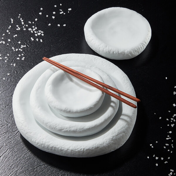 A set of white, round porcelain dishes with textured edges, stacked together with a pair of wooden chopsticks resting on top, displayed on a dark surface with scattered coarse salt.