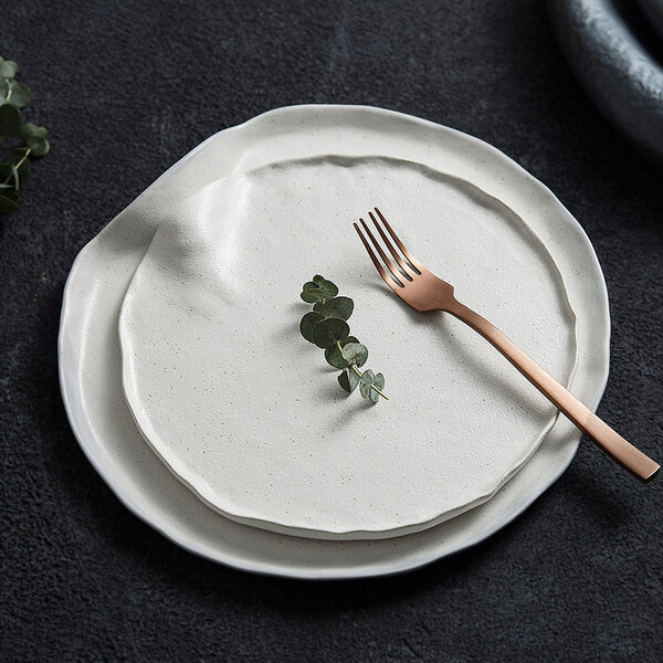 Two white, irregularly shaped stoneware plates are stacked on a dark textured surface, with a gold-colored fork and a small sprig of greenery placed on the top plate.