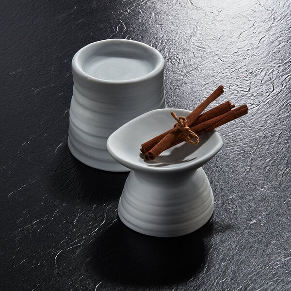 Two white porcelain dishes with a modern, wavy design are shown on a dark textured surface, one holding a bundle of cinnamon sticks.