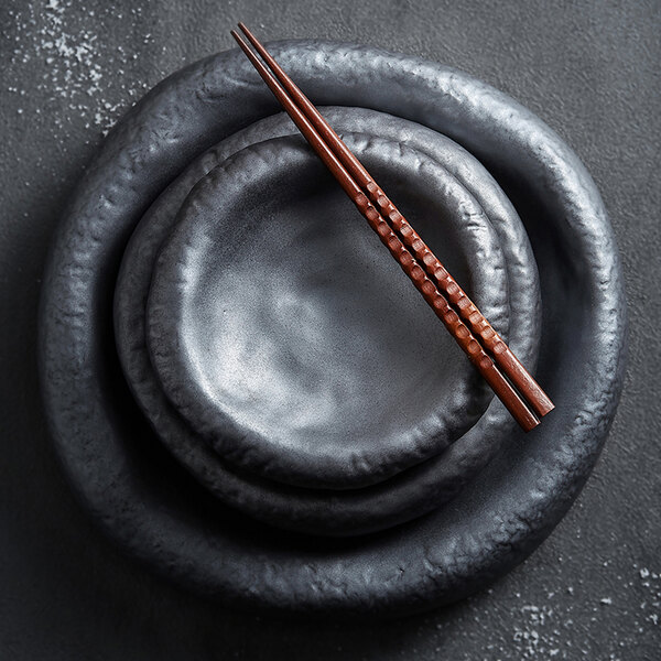 A set of three nested, round, matte black plates with a pair of brown chopsticks placed on top.