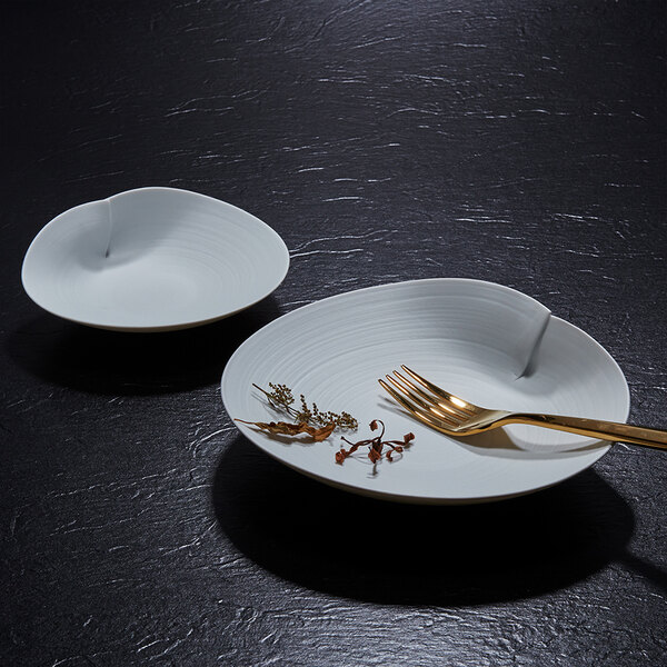 Two white, uniquely shaped porcelain plates are displayed on a dark textured surface, with one plate holding a gold fork and some dried herbs.