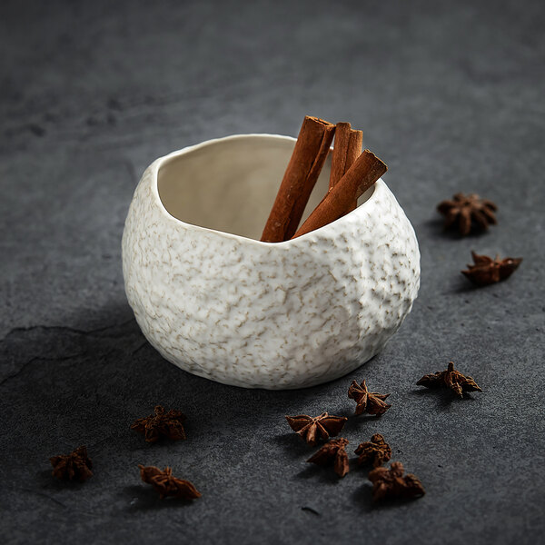 A textured white bowl containing cinnamon sticks, surrounded by star anise on a dark surface.