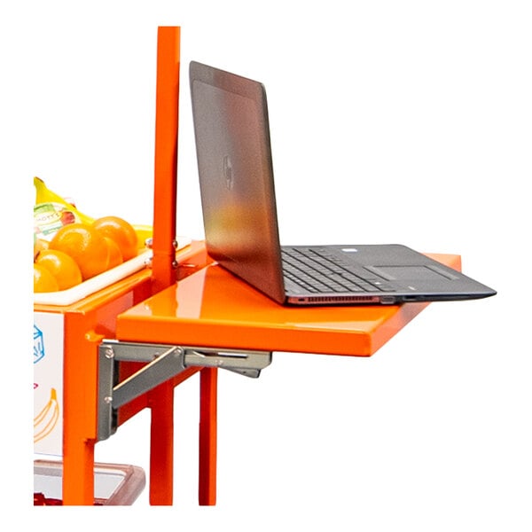 A black laptop is placed on an orange folding shelf attached to a metal frame, with some oranges visible in a container nearby.