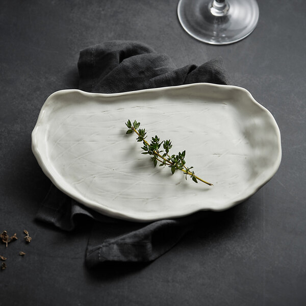 A white, irregularly shaped oval plate with a sprig of herbs placed on it, set on a dark cloth and surface with part of a wine glass visible in the background.