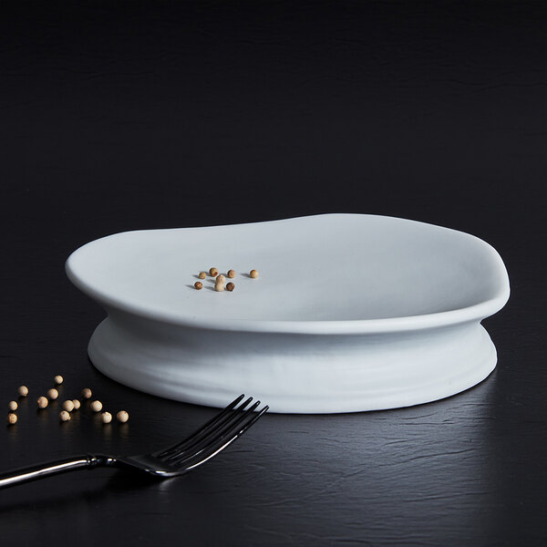 A white, round, slightly irregular stoneware plate with a raised base, shown with a few peppercorns and a fork nearby on a dark surface.