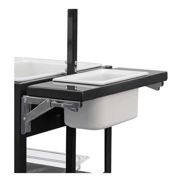 A partial view of a black metal cart with a foldable shelf holding a white rectangular pan.