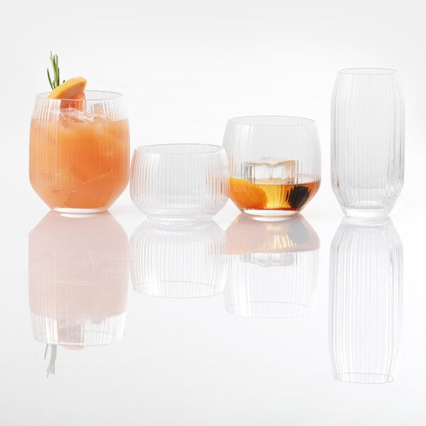 Four clear, ribbed glasses of varying shapes and sizes are arranged in a row, with two containing drinks and garnishes, and their reflections visible on the glossy surface below.