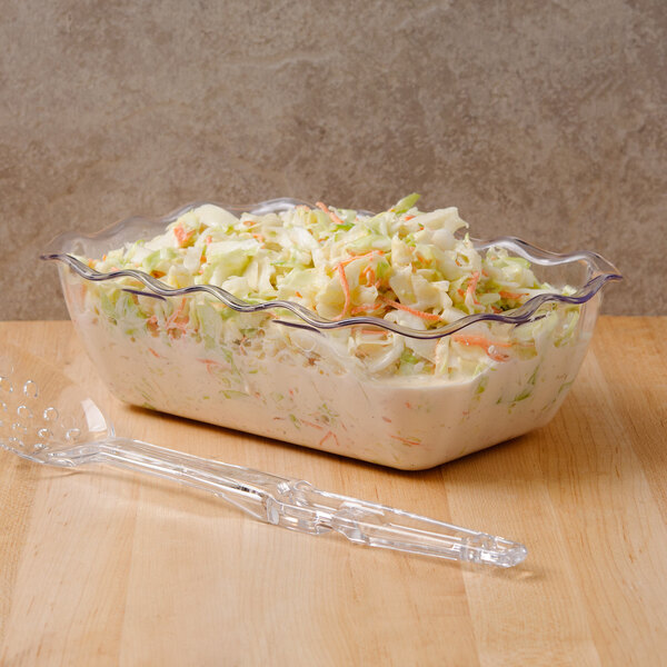 A clear, wavy-edged plastic container filled with creamy coleslaw, placed on a wooden surface next to a clear plastic serving utensil.