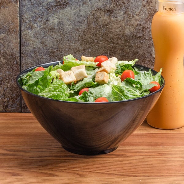 A black round bowl filled with salad greens, cherry tomatoes, and croutons, placed on a wooden surface next to a bottle of orange salad dressing.