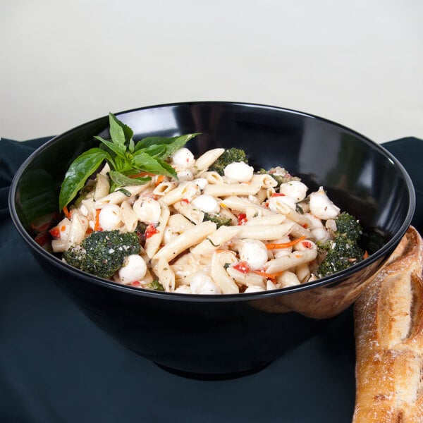 A black bowl filled with penne pasta, mozzarella balls, broccoli, red peppers, and garnished with fresh basil, with a piece of bread beside it.