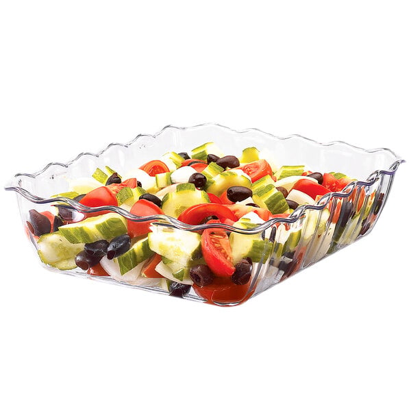 A clear, square, scalloped-edge plastic container filled with a colorful salad of cucumbers, tomatoes, onions, and black olives.