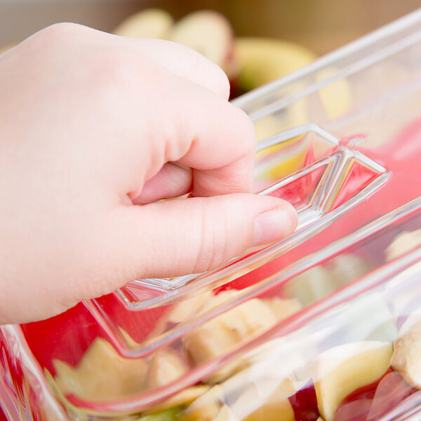 A hand is lifting a clear plastic lid with a rectangular handle from a container filled with cut fruit or vegetables.