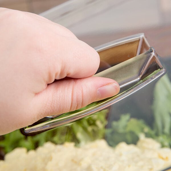 A hand is lifting a clear plastic lid with a handle, covering a container with food inside.