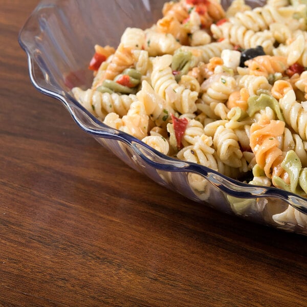 A clear, scalloped-edge bowl filled with tri-color rotini pasta salad on a wooden surface.