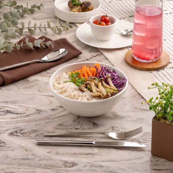 A white bowl filled with noodles, mushrooms, shredded carrots, chopped green onions, and shredded purple cabbage is placed on a marble table alongside a fork, knife, and other tableware.