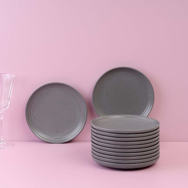 A stack of gray round plates with two additional plates standing upright, set against a pink background with a clear glass on the left.