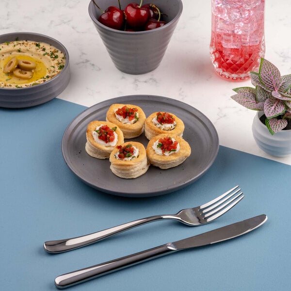 A gray round plate with five small pastry appetizers, placed on a blue placemat with a fork and knife nearby; other items include a bowl of cherries, a dish of hummus with olives, a pink drink, and a small potted plant.