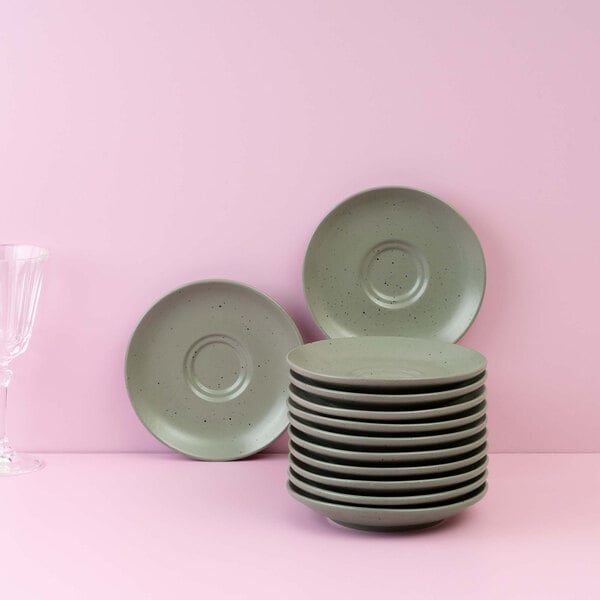 A stack of round, green saucers with two additional saucers propped up behind them, set against a pink background with a clear glass on the left.