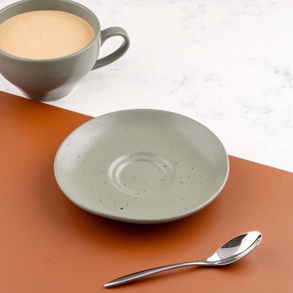 A round, light green saucer with a speckled pattern is placed on a brown surface, accompanied by a matching cup of tea or coffee and a shiny metal spoon.