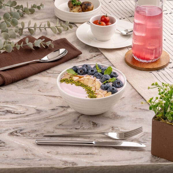 A round white bowl filled with a pink smoothie topped with blueberries, sliced almonds, green garnish, and mint leaves, placed on a marble-patterned table with cutlery, a glass of pink drink, and other dishes nearby.