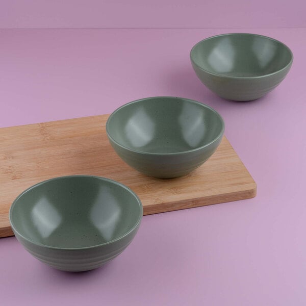 Three green bowls are arranged on and beside a wooden cutting board against a pink background.