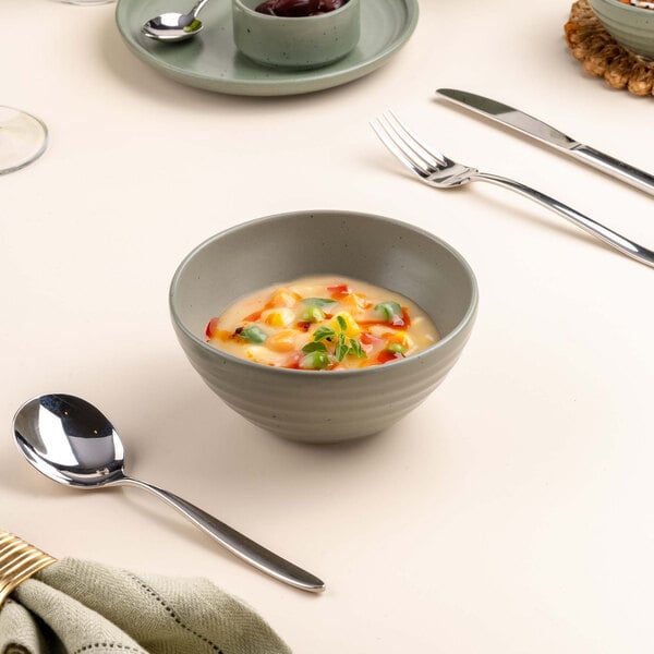 A small green bowl filled with a creamy soup garnished with herbs and diced vegetables, placed on a set dining table with cutlery and a spoon nearby.