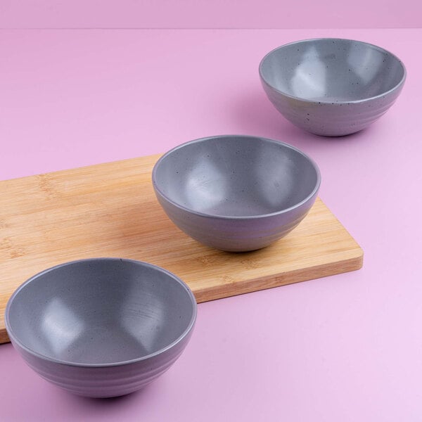 Three gray round bowls are arranged on and beside a wooden cutting board against a pink background.