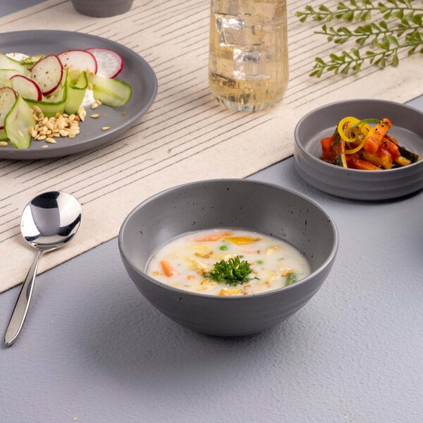 A gray round bowl filled with creamy soup garnished with parsley, placed on a table alongside a spoon, a plate of salad, a small dish of vegetables, and a glass of beverage.