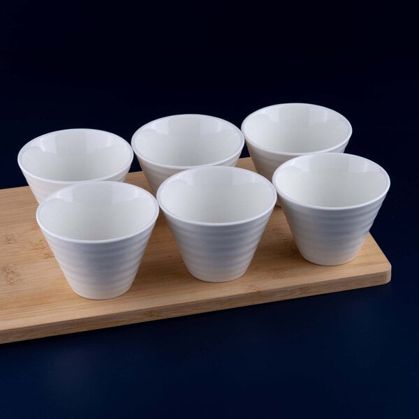 Six white, round porcelain bowls are arranged on a rectangular wooden board against a dark background.