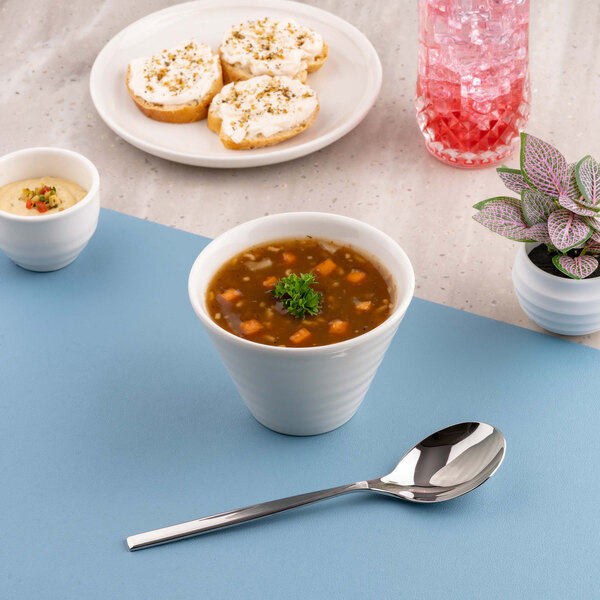 A white porcelain bowl filled with soup, garnished with herbs, is placed on a blue surface alongside a spoon, with other food items and a plant visible in the background.