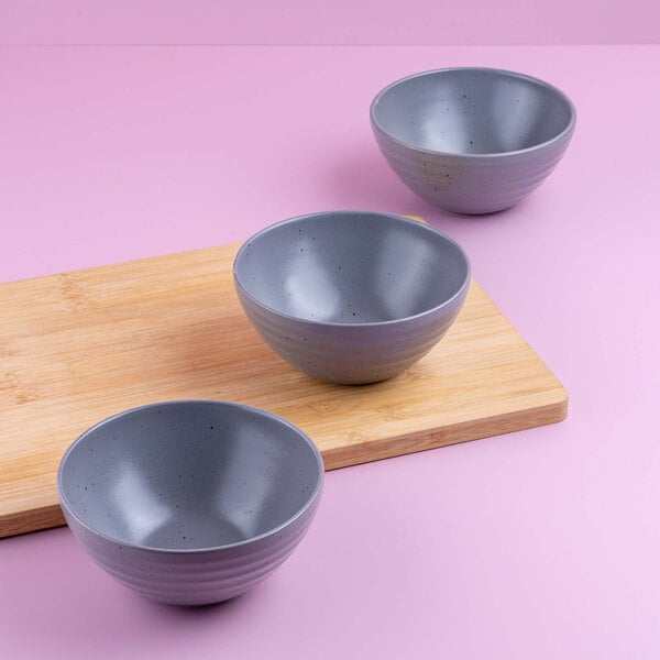 Three gray round bowls are arranged on and around a wooden cutting board against a pink background.