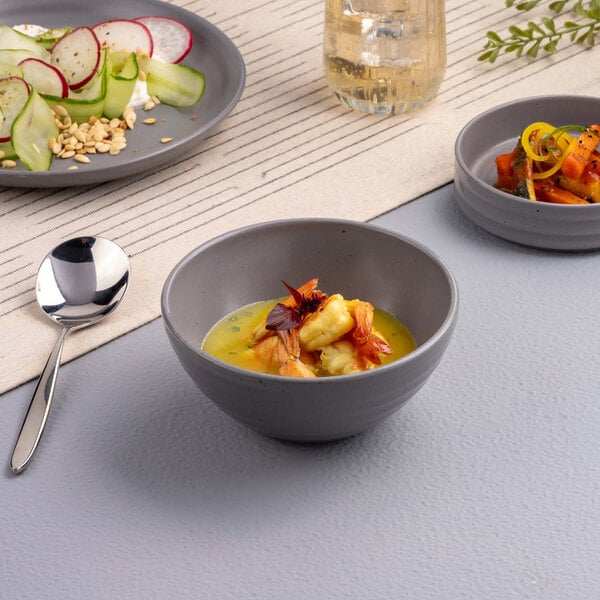 A small gray bowl filled with a yellow soup or sauce and garnished with pieces of seafood and herbs, placed on a light-colored surface with other dishes and a spoon nearby.