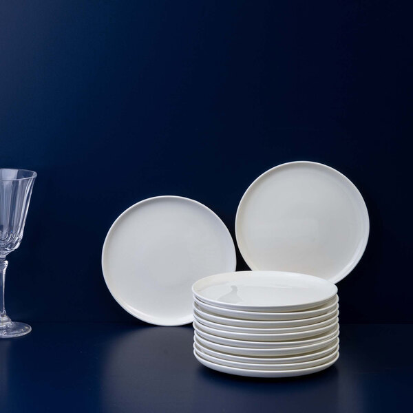 A stack of twelve plain white round plates is displayed on a dark surface with two plates standing upright behind the stack and a clear wine glass to the left.
