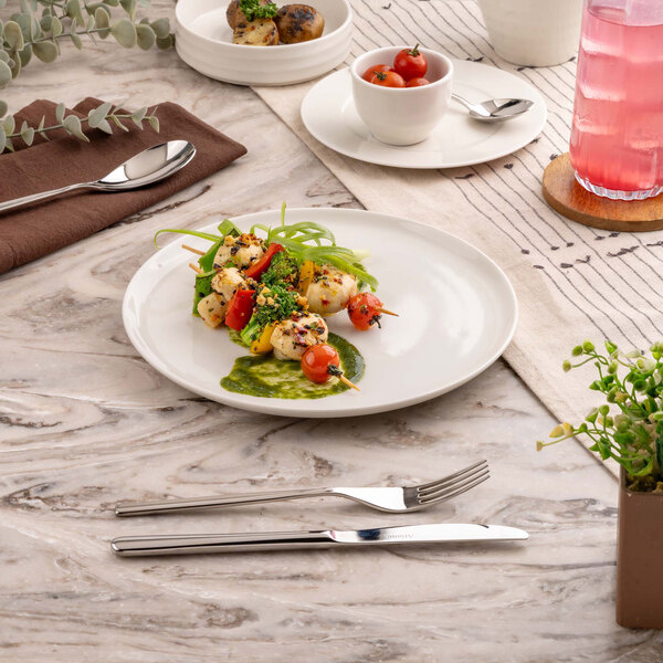 A round white plate with a colorful vegetable and herb dish, set on a marble table with cutlery, a napkin, and a glass of pink beverage nearby.