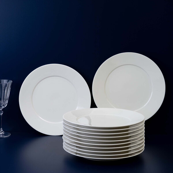A stack of plain white round plates is displayed in front of two upright plates against a dark background, with a glass visible on the left side.