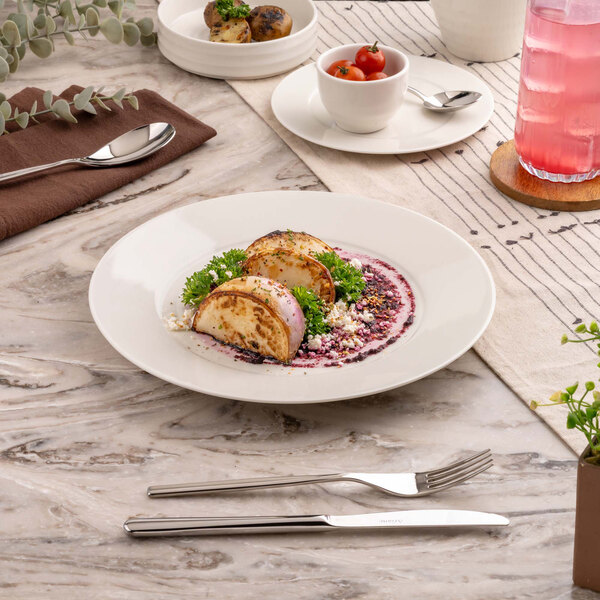 A round white plate with a gourmet dish featuring two pieces of grilled or roasted vegetables, garnished with greens and a colorful sauce, set on a marble table with cutlery and other tableware nearby.