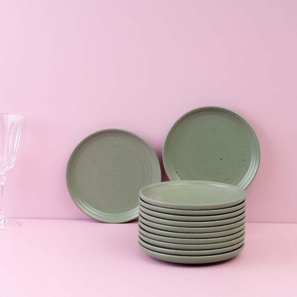 A stack of round, light green plates with two additional plates standing upright in the background, set against a pink surface and wall, with a clear glass partially visible on the left.