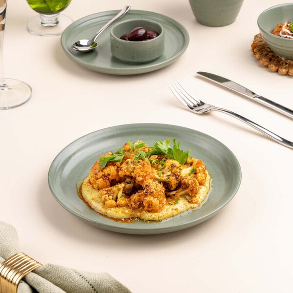 A green ceramic plate with a serving of roasted cauliflower garnished with herbs, placed on a light-colored table with cutlery and other dishes nearby.