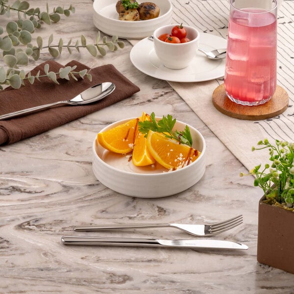 A round white bowl filled with orange slices and garnished with herbs, placed on a marble surface alongside cutlery, a glass of pink beverage, and other tableware.