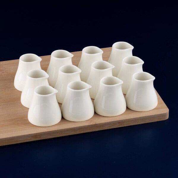 Twelve small white porcelain creamers are arranged on a wooden board against a dark background.